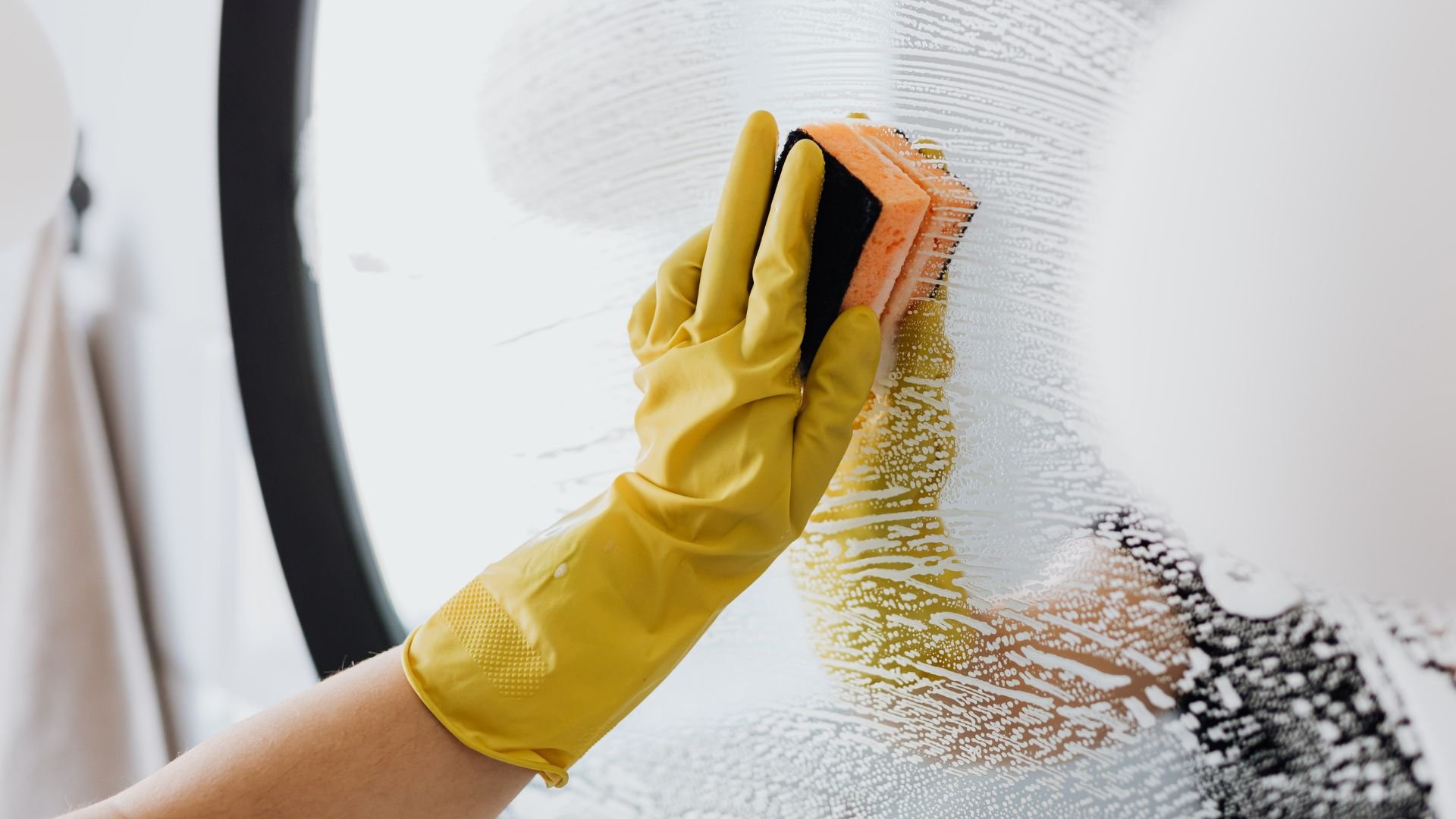 Yellow gloved hand scrubbing white surface with orange sponge